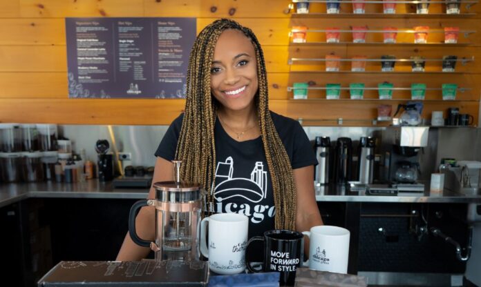 Kris Christian of Chicago French Press smiles behind a display of coffee products, including French presses, mugs, and specialty coffee bags in a modern cafe setting.
